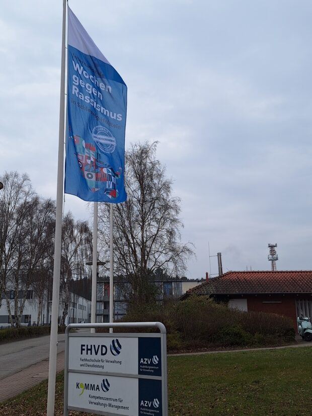Eine blaue Fahne mit der Aufschrift "Wochen gegen Rassismus" weht neben einem Schild des FHVD und anderer Organisationen vor einem Gebäude mit Bäumen und einem bewölkten Himmel im Hintergrund.