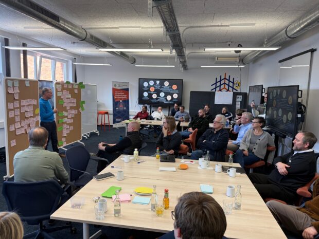 Eine Gruppe von Personen sitzt in einem Besprechungsraum um Tische herum und hört einem Vortragenden zu, der an einer mit Haftnotizen bedeckten Tafel steht. Laptops, Papiere und Getränke stehen auf den Tischen. Es herrscht eine kollaborative Atmosphäre.