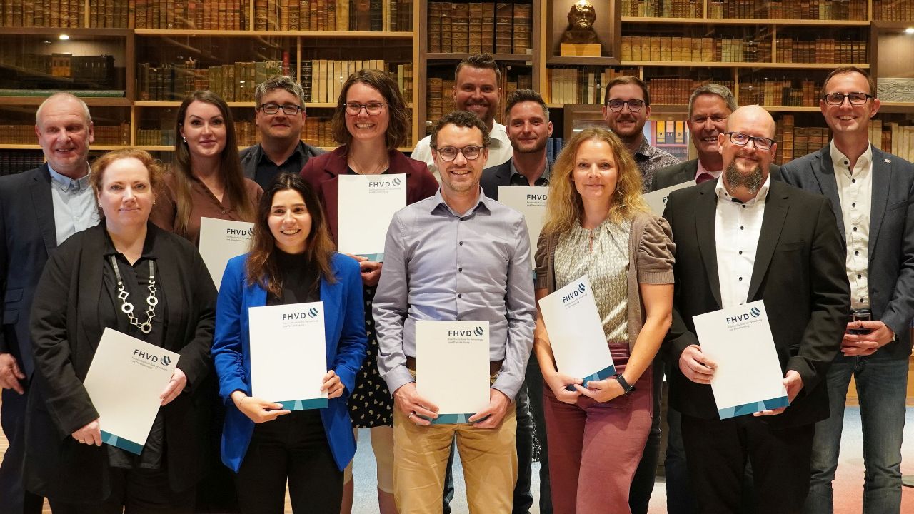 Eine Gruppe von fünfzehn Personen steht in einem Raum, lächelt und hält Urkunden in der Hand. Im Hintergrund sind mit Büchern gefüllte Regale zu sehen, die eine formale, akademische Atmosphäre schaffen.