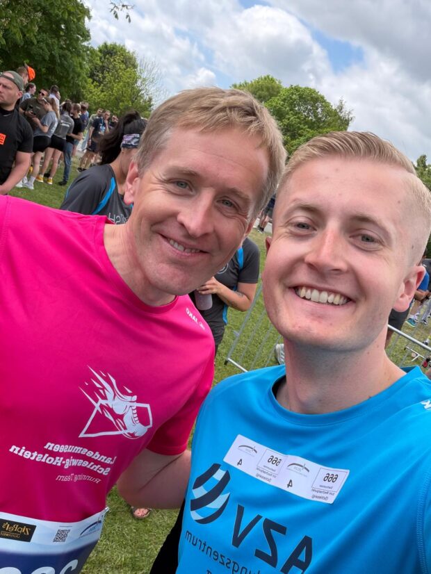 Zwei lächelnde Männer machen bei einem Laufevent im Freien ein Selfie. Der eine trägt ein rosa T-Shirt, der andere ein blaues T-Shirt, an denen Startnummern befestigt sind. Im Hintergrund sind Menschen, Bäume und ein bewölkter Himmel zu sehen.