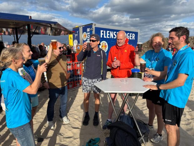 Eine Gruppe von Menschen steht an einem Sandstrand, lächelt und stößt gemeinsam an einem kleinen Tisch mit ihren Getränken an. Einige tragen Sportkleidung und Medaillen, was darauf hindeutet, dass sie gerade ein Rennen beendet haben. Der Himmel ist teilweise bewölkt. Im Hintergrund steht ein blauer Wohnwagen.