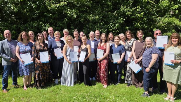 Eine Gruppe von etwa 25 Personen steht draußen auf einer Wiese, lächelt und hält Urkunden in den Händen. Im Hintergrund sind Bäume und Grünflächen zu sehen, und die Gruppe scheint an einem sonnigen Tag einen Erfolg zu feiern.