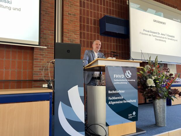 Ein Mann im Anzug spricht mit einem Mikrofon am Rednerpult. Hinter ihm wird eine Präsentation gezeigt. Auf der Bühne steht ein Blumenarrangement, und auf einem Schild steht „FHVD – Fachhochschule für Verwaltung und Dienstleistung“.